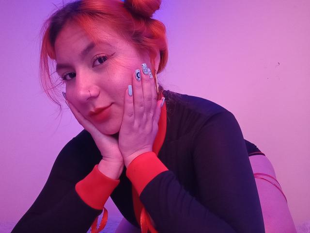 AradiaX with orange hair in buns, black and red long-sleeve top, blue nails, hands on face, smiling, pink and purple lighting, visible butt in red underwear.