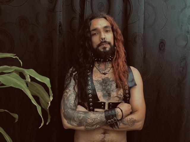 EscariotNEeva, avec de longs cheveux roux, des tatouages, un harnais en cuir noir, des manchettes, un anneau de nez et une barbe, debout devant des rideaux sombres, les bras croisés, avec une plante verte sur la gauche..