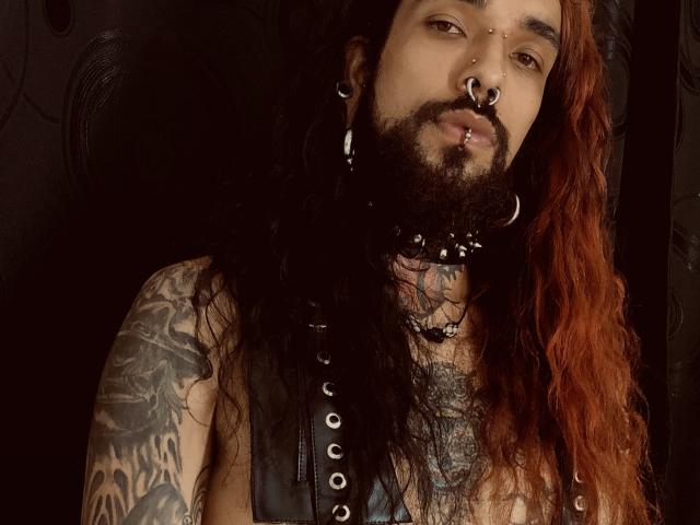EscariotNEeva, con cabello rojo largo, barba, múltiples perforaciones en la nariz, perforación en el labio, collarín negro, brazos tatuados y una manga negra, mirando a la cámara. Fondo oscuro..
