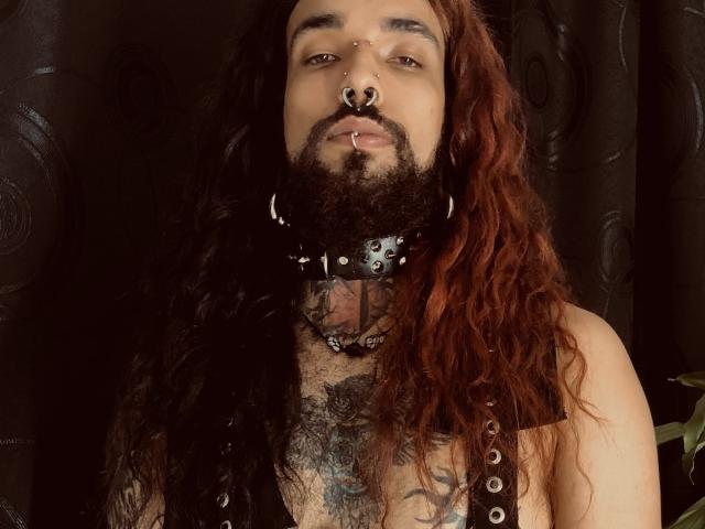 EscariotNEeva, con lunghi capelli rossi ricci, barba, anello al naso, anello al labbro, collare di pelle nera con borchie metalliche, tatuaggio sul petto e orecchini a cerchio, guarda verso l'alto con un'espressione seria..