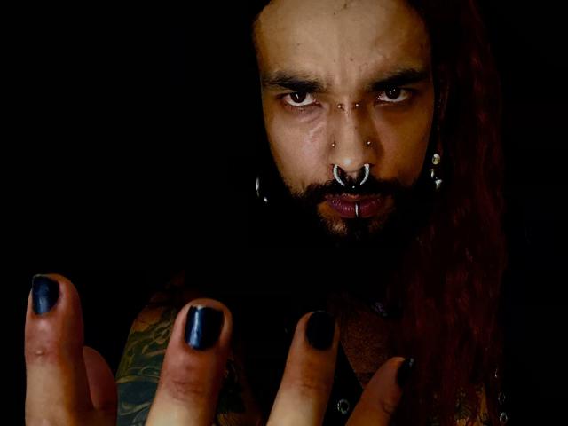 EscariotNEeva, com dreadlocks vermelho-escuro, esmalte de unha preto, piercing no nariz, piercing no lábio e tatuagens, olhando intensamente para a câmera com uma expressão séria, com as mãos levantadas na frente..