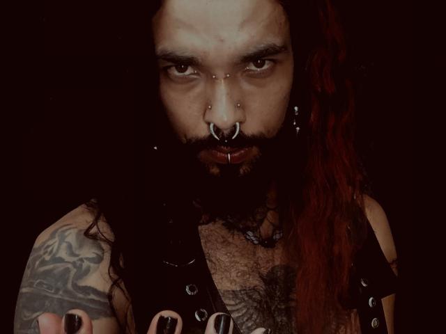 EscariotNEeva con rastas rojas, piercings en la nariz y los labios, chaleco de cuero negro sin mangas, tatuajes y una expresión seria. Fondo oscuro. Vello facial abundante. Piel ligeramente sucia..