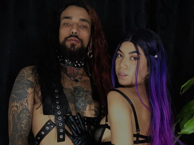 EscariotNEeva, con i capelli rosso scuro, la barba, il piercing al naso, un'imbracatura in pelle nera e tatuaggi, è in piedi accanto a EscariotNEeva, con i capelli viola, un reggiseno e mutandine neri a spalline e uno sguardo di lato..