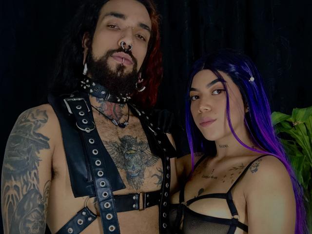 EscariotNEeva, con lunghi capelli viola e tatuaggi, indossa un intimo nero con spalline sottili, e si trova accanto a un uomo barbuto con tatuaggi, capelli rossi e neri, e un'imbracatura in pelle nera..