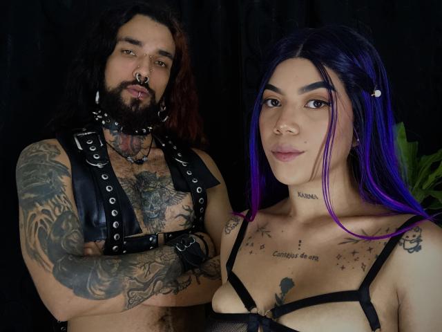 EscariotNEeva, con i capelli viola, piccoli seni e tatuaggi, è in piedi accanto a un uomo tatuato e con la barba, con i capelli lunghi, un'imbracatura di pelle nera e un pene grande e flaccido. Entrambi hanno uno sfondo nero..
