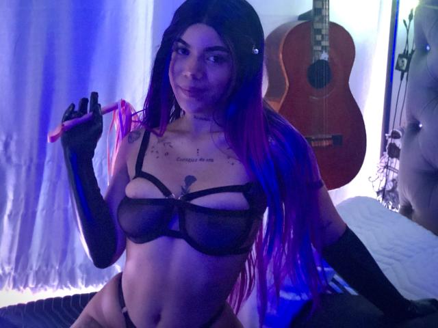 EscariotNEeva, con capelli viola e blu, indossa un reggiseno e dei guanti di rete neri, tiene un nastro rosa, mostra i seni, in una stanza con una chitarra e un letto bianco. Sorride, sembra carina..