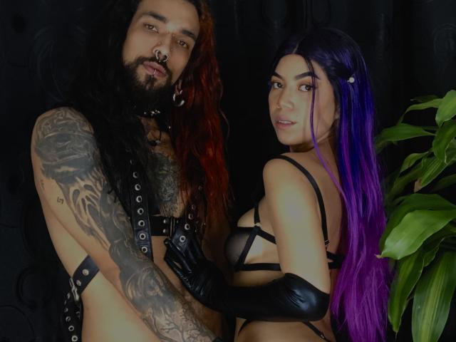 EscariotNEeva, avec de longs cheveux roux et noirs, des tatouages et un harnais en cuir noir, se tient à côté d'EscariotNEeva, qui a les cheveux violets, un soutien-gorge noir et des gants, et les deux regardent l'appareil photo..