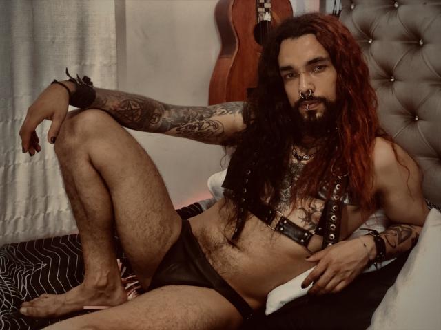 EscariotNEeva, com cabelos ruivos longos, tatuagens e barba, vestindo uma coleira de couro preta e roupa íntima, sentada em uma cama com as pernas abertas, exibindo um corpo musculoso e peludo..