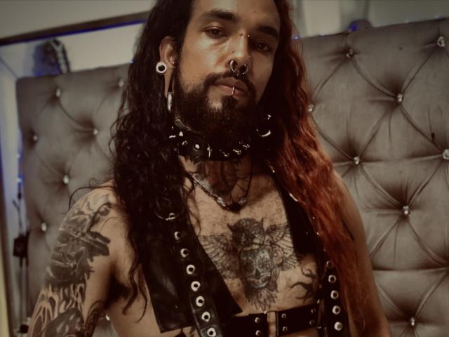EscariotNEeva with long red-black hair, beard, nose ring, and ear gauges, wearing black leather harness with metal studs, tattoos on chest and arm, serious expression, indoors.