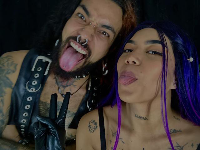EscariotNEeva mit lila Haaren, die die Zunge herausstreckt, neben EscariotNEeva mit einer schwarzen Lederweste, Bart, Nasenring und herausgestreckter Zunge, beide zeigen Tattoos und machen "Rock on"-Handzeichen..