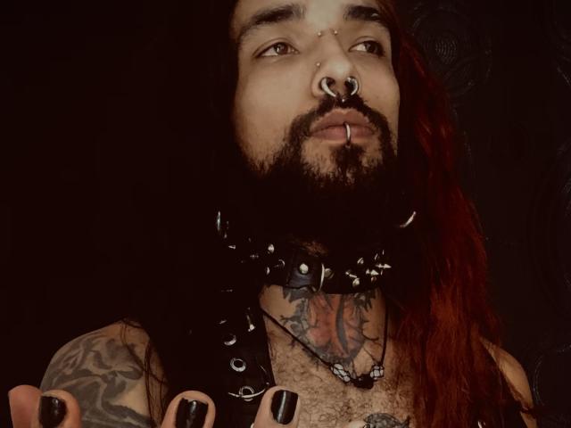 EscariotNEeva, avec des cheveux roux, des tatouages sur la poitrine et le bras, un collier en cuir noir, du vernis à ongles noir, des piercings au nez et aux lèvres, regardant vers le haut avec une expression sérieuse..
