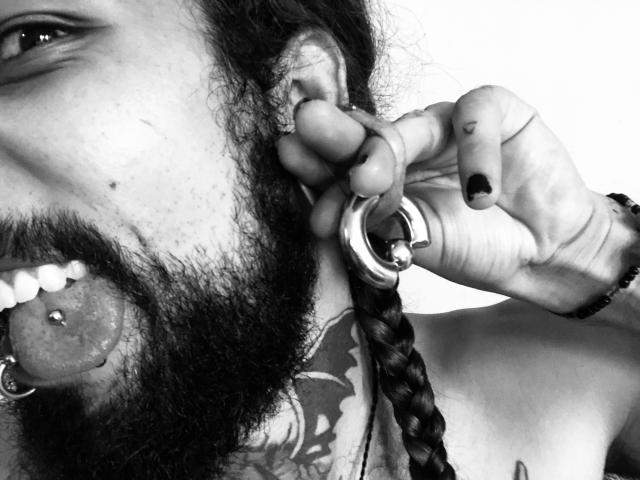 EscariotNEeva, com uma barba espessa e cabelo trançado, com a língua perfurada para fora, segurando um anel com a mão de unhas pretas, mostrando uma tatuagem no pescoço..