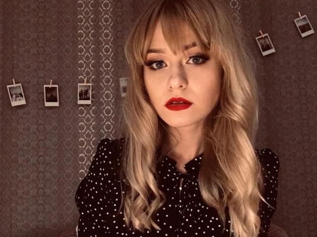 CindyxGlam has long blonde hair, red lipstick, and black polka-dot dress, with large tits and sultry eyes, hanging polaroid pics in the background.