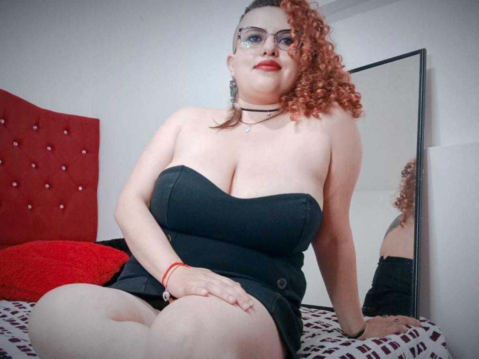 Hanna Harendt, avec ses cheveux roux bouclés, des lunettes, une robe noire sans bretelles, montrant sa poitrine généreuse, assise sur un lit, avec une tête de lit rouge, un miroir, un oreiller rouge, un mur blanc, un bracelet rouge..