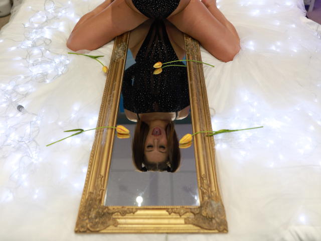 MandyPeas is kneeling, legs spread, wearing a black sparkly dress, reflected in an ornate mirror, with tulips and fairy lights on a white bed. Her mouth is open.