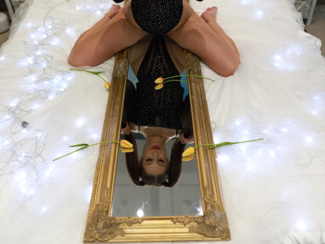 MandyPeas is on her back with legs spread, wearing a sparkly black dress, reflected in a gold mirror, with yellow tulips and white lights around her.