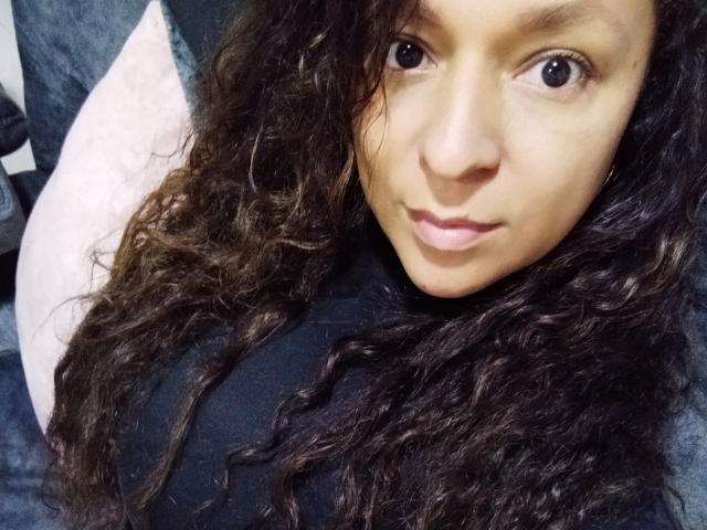 Angelonix with curly brown hair, medium skin, and brown eyes, wearing a black top, has a small nose and pink lips, looking directly at the camera with a slight smile.