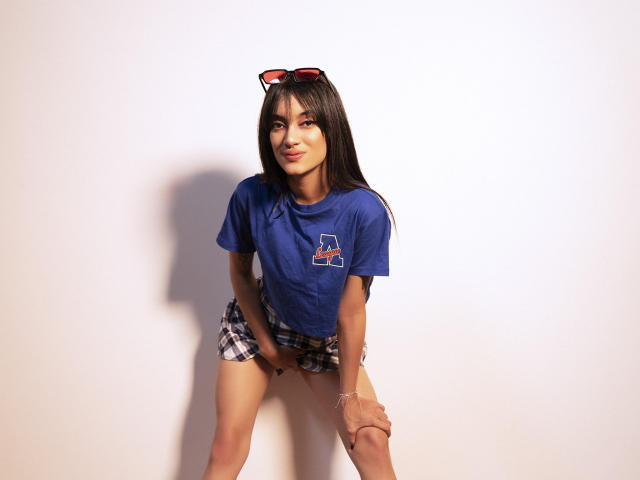 MeliMuller with long black hair, blue t-shirt, plaid shorts, red sunglasses on head, smiling, standing, hands on knees, shadow on white wall, visible tattoo on right thigh.