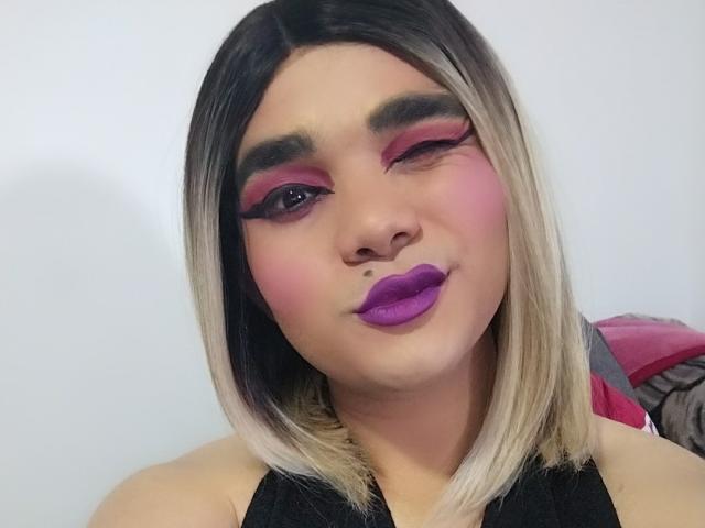 SexyHottieTS met een kort zwart en blond kapsel, knippend, roze en paarse oogmake-up, opvallende paarse lippenstift, draagt een zwarte top, witte achtergrond..