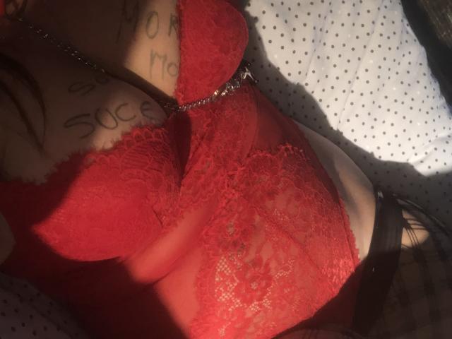 A lingerie vermelha de renda da SerenaFr, mostrando seus seios grandes e uma tatuagem no peito, com alças pretas e uma corrente brilhante, em pouca luz..