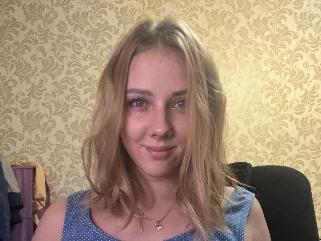 LaryAngelMy a les cheveux blonds et ondulés, des yeux bleus et un sourire charmant. Elle porte un haut bleu à pois et un collier délicat avec un petit pendentif..