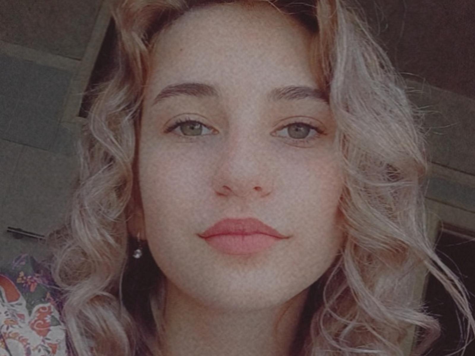 JennyLizzie has blonde, wavy hair, green eyes, and pink lips. She's wearing a floral shirt, and her small, round, pink nipples are barely visible under her shirt.