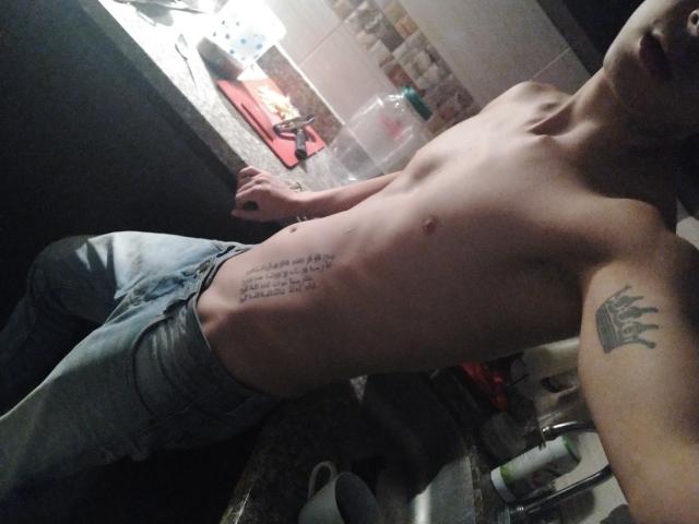 LatinAndres, sem camisa, deitado na bancada da cozinha, com tatuagem visível na lateral, tatuagem no braço, vestindo jeans, com uma leve barriga, tom de pele médio, fundo escuro, relaxado, selfie com um leve ângulo..