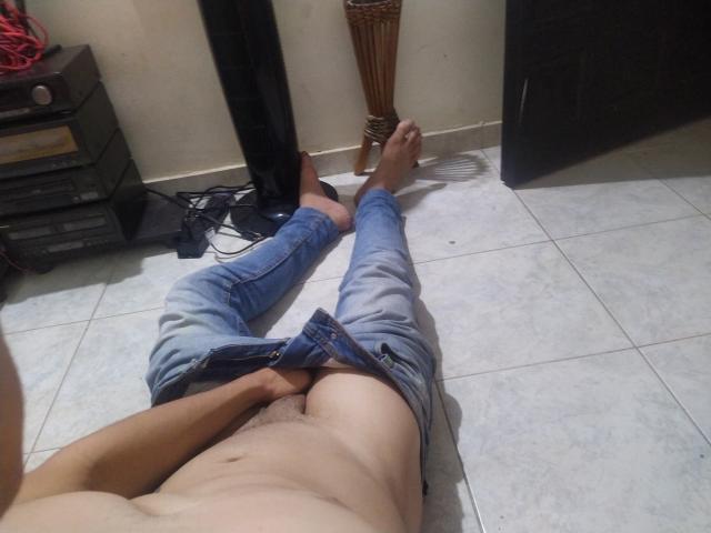 LatinAndres, sem camisa, com o peito descoberto e o pênis visível, deitado no chão de azulejo, pernas abertas, calças jeans azuis desabotoadas, mão cobrindo a virilha, sapatos pretos e cabos à esquerda..
