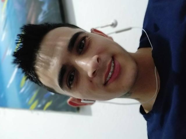 LatinAndres tem um cabelo preto curto e espetado, olhos castanhos e um pequeno sorriso. Ele está vestindo uma camisa azul escura com um colar de prata e fones de ouvido vermelhos..