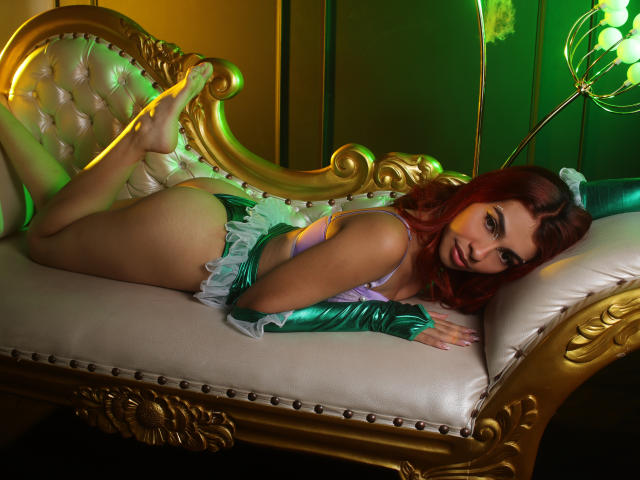 LeahMarie, with red hair, lies on a golden, tufted couch, wearing green and white lingerie, showing her ass and tits, with green and yellow lighting.