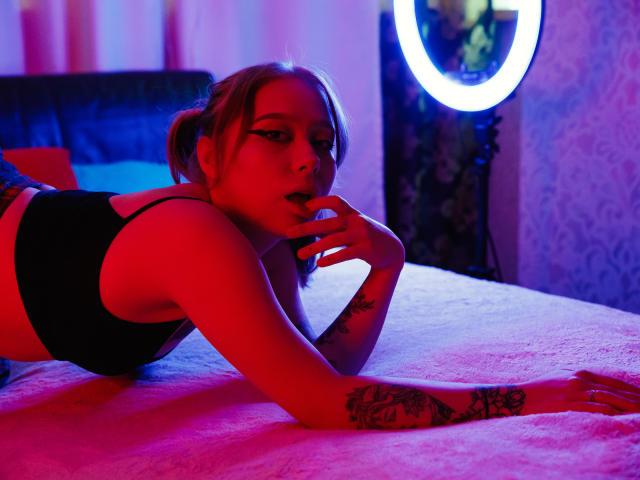 Hayley Trent, con cabello rubio, sostén negro, acostada en la cama, iluminación roja y azul, dedo en la boca, tatuajes en el brazo, expresión seductora, sábana rosa, luz de aro en el fondo..