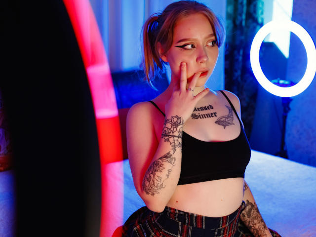 Hayley Trent mit hochgezogenen Lippen, Hand am Kinn, schwarzes Crop-Top, karierter Rock, Tattoos am Arm und an der Brust, Neon-Ringlicht, rosa und blaue Lichter, blonde Zöpfe..