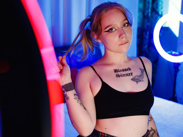 HayleyTrent with pigtails, black crop top, tattoos, and eyeliner, holding a ponytail, neon lights, ring light, and blue curtains in the background. Her tits are small and cute.