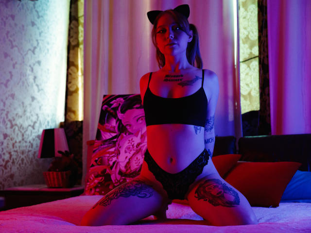 HayleyTrent with cat ears, black bra, and lace panties, kneeling on a bed, showing tattoos on thighs and chest, under purple and blue lighting.