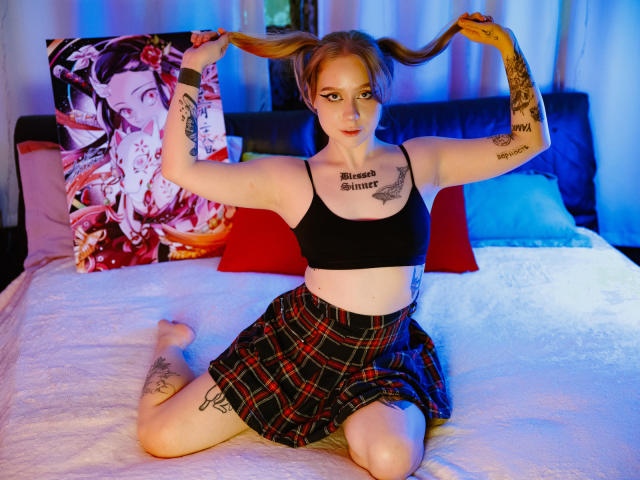 Hayley Trent, con coletas, un top deportivo negro y una falda escocesa roja, está arrodillada en la cama, mostrando tatuajes, su trasero desnudo y tatuajes en los muslos, en una habitación colorida, sosteniendo las coletas hacia arriba..
