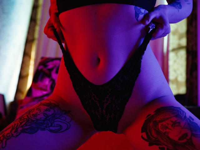 Hayley Trent se está quitando unos panties de encaje negros, revelando su vulva. Tiene tatuajes en los muslos y un ombligo visible. La iluminación es azul y rosa..