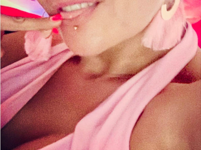 La robe rose de KiwiJuicy, qui dévoile sa poitrine et un petit piercing au mamelon. Elle mord sa petite doigt rose manucuré, et porte des boucles d'oreilles en plumes roses ainsi qu'un sourire discret..