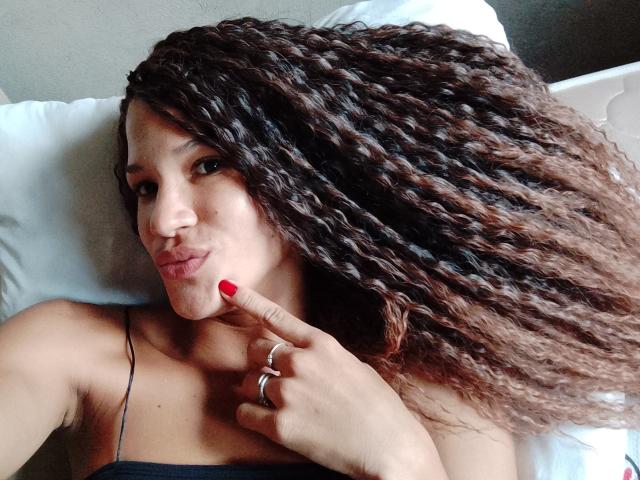 Paulina Nicolle, avec ses cheveux bruns bouclés, ses ongles rouges et une bague, faisant une moue en touchant ses lèvres avec son doigt, allongée sur un oreiller blanc..