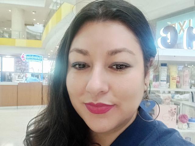 SakotoDika ha i capelli scuri e ondulati, occhi marroni, rossetto rosa e indossa una camicia blu con orecchini a cerchio, guardando leggermente di lato con un sorriso appena accennato..