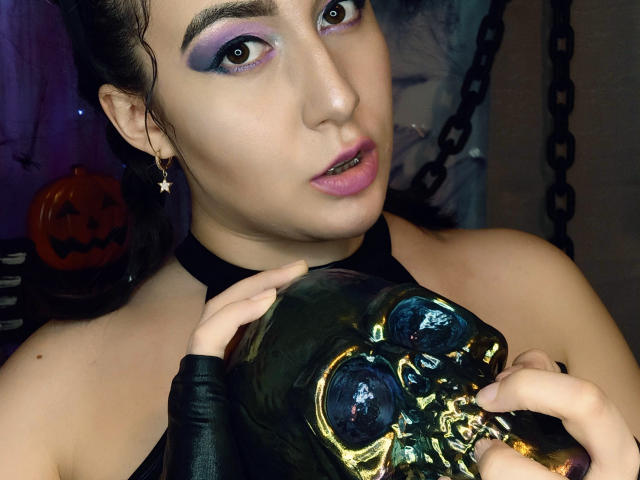MabyLegrand with purple eyeshadow, black choker, and star earrings, holding a shiny black skull, wearing black leather glove, pink lipstick, and looking at camera.