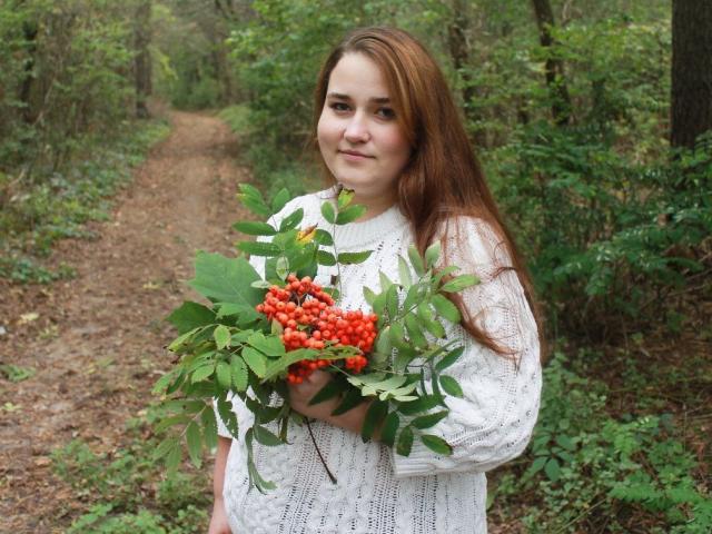 DaniellaGraceZZ with red hair, holding a bouquet of red berries and green leaves, wearing a white knit sweater, standing on a forest path, smiling slightly.