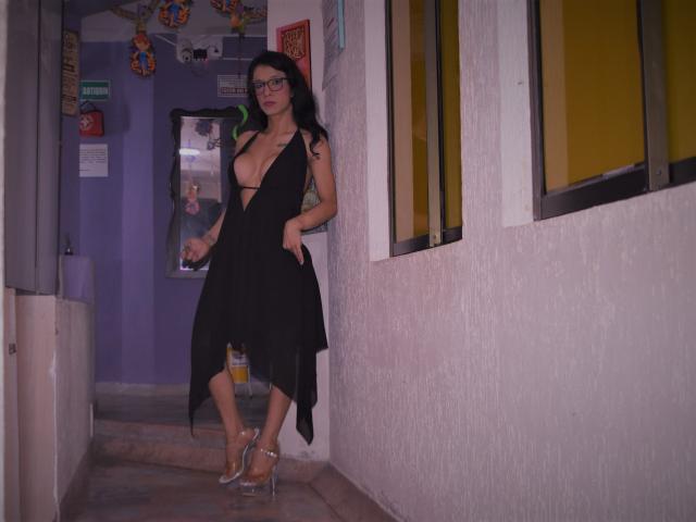Daslyroze69, avec de grandes seins, portant une robe noire avec un décolleté plongeant, debout dans un couloir, tenant une boisson, chaussée de talons transparents, a les cheveux foncés et porte des lunettes..