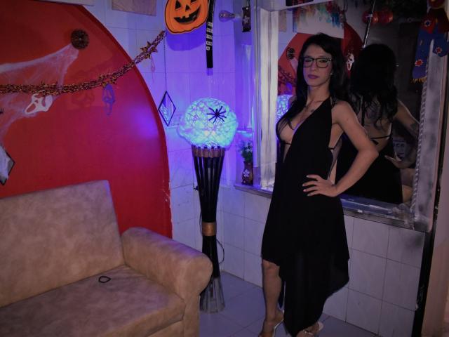 Daslyroze69, una mujer delgada con senos grandes y cabello oscuro, lleva un vestido negro con aberturas laterales, gafas y tacones blancos, y está de pie en una habitación decorada para Halloween..