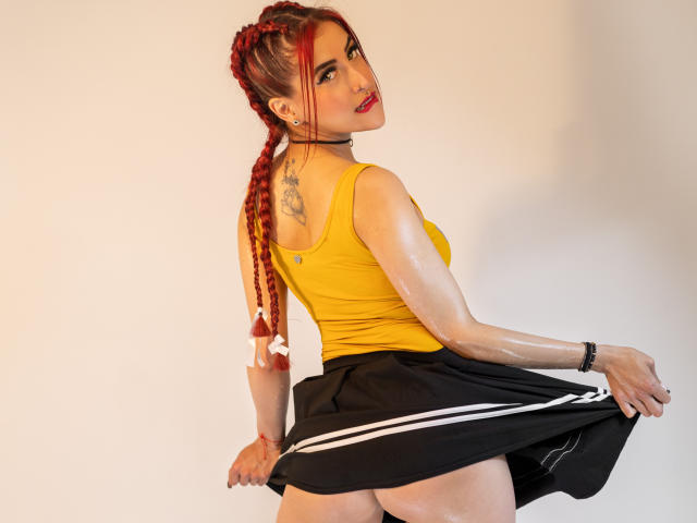 AlissaBrown with red braids, yellow top, black skirt, exposed shiny ass, tattoo on upper back, looking over shoulder, wearing glasses, white ribbon in hair.