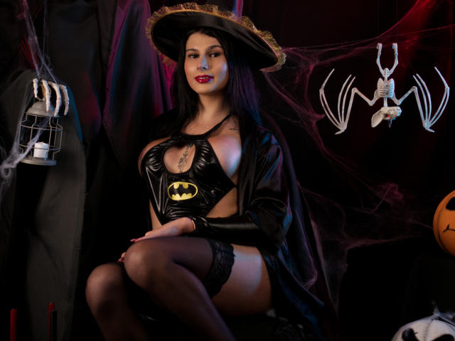 Maddymayer con un costume da Batman in pelle nera, che mostra il seno attraverso un'apertura, calze nere alte fino alla coscia e un mantello, seduta con un braccio tatuato, in un'atmosfera spaventosa di Halloween..