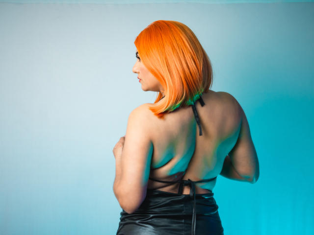 NatalyCollin with orange bob, back to camera, topless, black leather skirt, showing her ass, blue-green light on right, holding her tits with her arms.