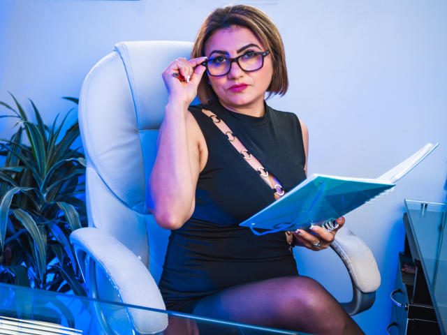NatalyCollin with glasses, black dress, and short brown bob, sitting in white chair, holding paper, adjusting glasses, blue lighting, glass table, plant in background, sexy, confident.