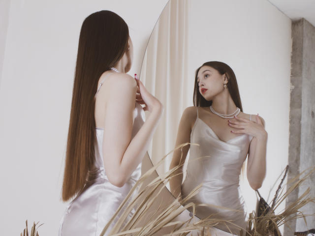 EllyCat with long brown hair in a shiny white satin dress, looking at her reflection, touching her chest, wearing a pearl necklace, standing in front of a mirror.
