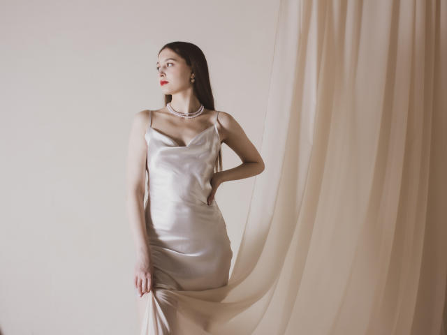 EllyCat with long dark hair, wearing a silky silver dress, red lipstick, and a pearl necklace, holding the dress's slit, standing next to flowing beige curtains.
