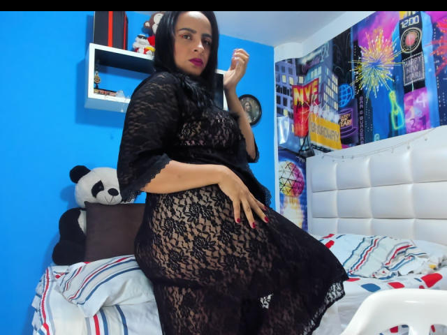 PamelaRouss, vestindo um vestido preto com detalhes em renda, está sentada na cama, mostrando seus seios e nádegas, com batom vermelho escuro, cabelo preto, parede branca, parede azul, cabeceira branca e um brinquedo de pelúcia em formato de panda..